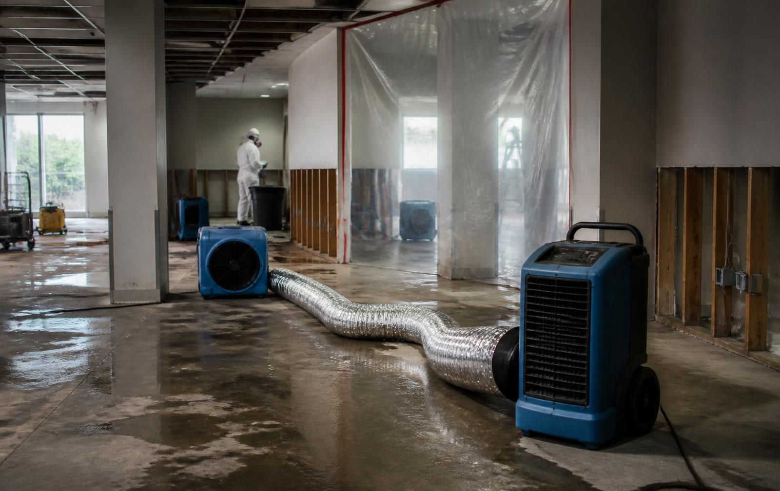 Dehumidifiers, an air mover, and flex ducting running across a wet concrete floor beside plastic containment sheeting in a commercial building stripped to the studs during water damage restoration