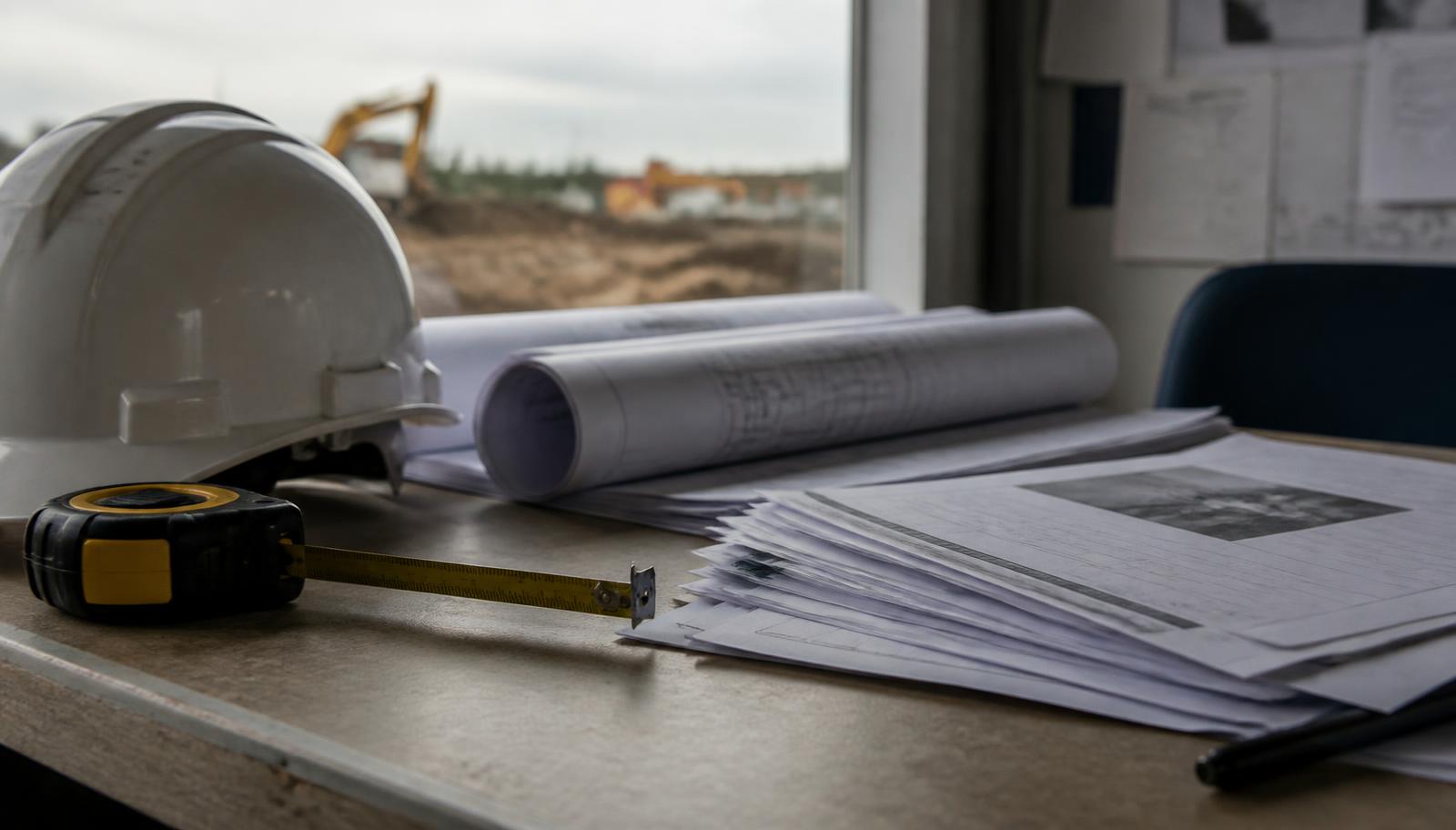 Rolled drawings, a hard hat, a tape measure, and a stack of printed project documents on a site trailer table beside a window overlooking a graded site