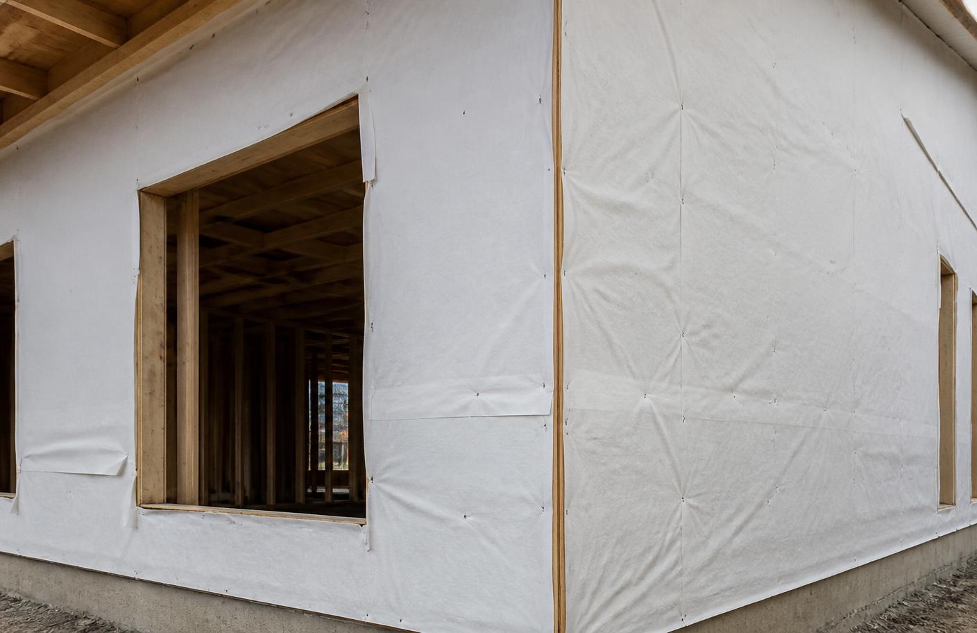 House wrap stapled over the wood framing of a tract home, with an open window rough opening showing the joists inside